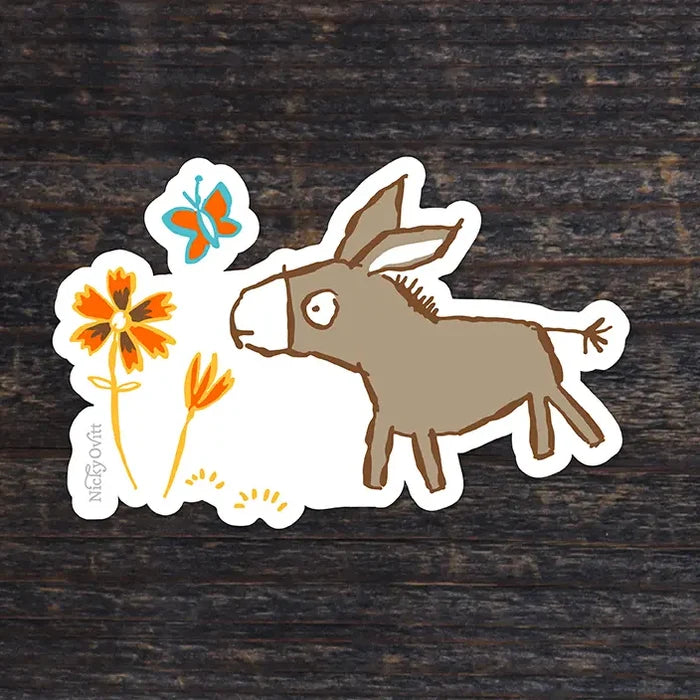 A die-cut sticker featuring a whimsical illustration of a brown donkey, orange flowers, and a blue butterfly on a white background, placed on a dark wood surface. The brand name 'Nico D'Or' is printed vertically on the left edge of the sticker.