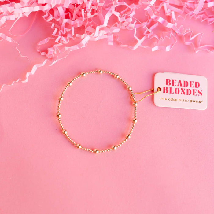 A gold-colored bracelet with beads, displayed on a pink background with crumpled pink paper and a tag that reads 'BEADED BLONDES'.