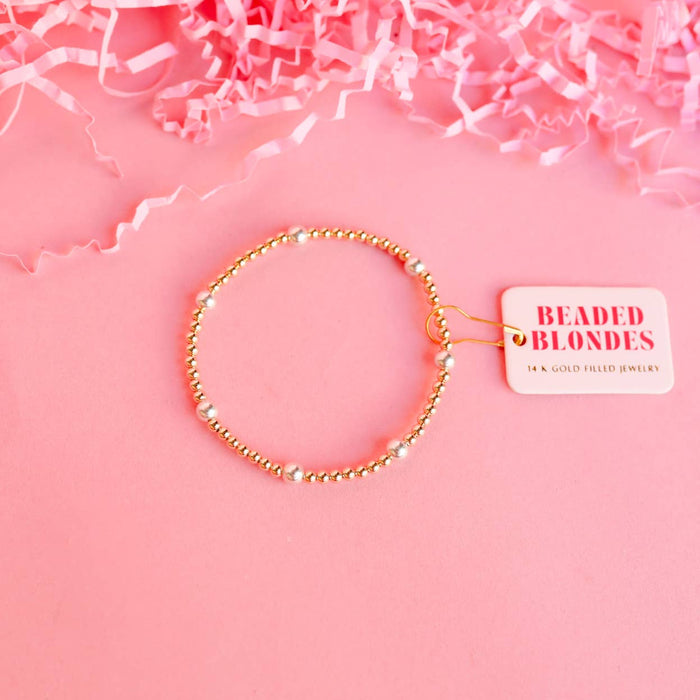 A gold-colored bracelet with silver beads displayed on a pink background with crumpled pink paper and a tag that reads 'BEADED BLONDES'.