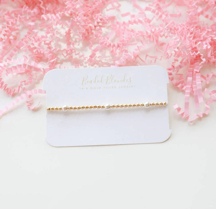 A gold-colored bracelet with silver beads displayed on a pink background with crumpled pink paper and a tag that reads 'BEADED BLONDES'.
