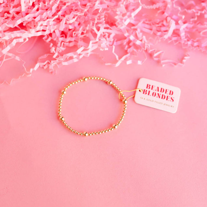 A gold-colored bracelet made of filled beads, displayed on a pink background with crinkled pink paper to the side and a tag attached to the bracelet.