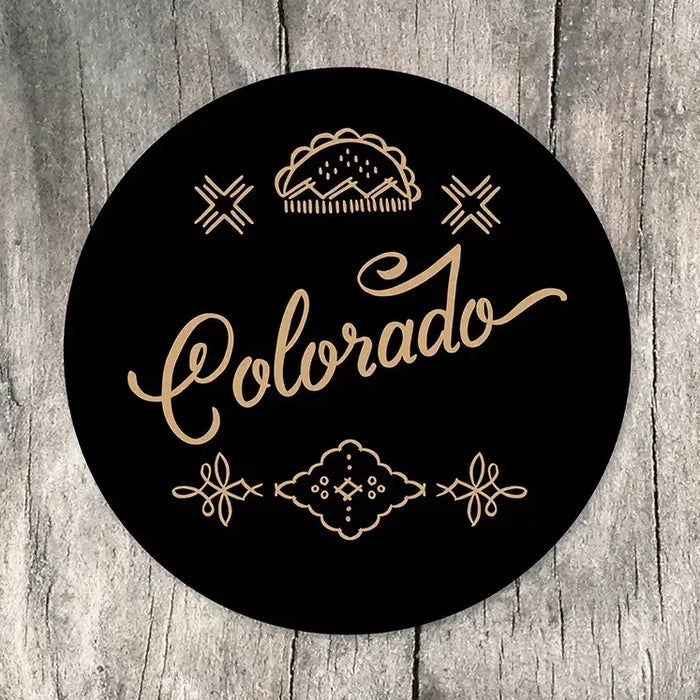 A round black sticker featuring the word 'Colorado' in elegant gold cursive script, surrounded by decorative gold line art including a mountain silhouette, stars, and ornamental flourishes, displayed on a rustic gray wood background.